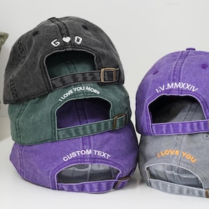 May include: A stack of six baseball caps in various colors, including black, green, purple, and gray. The caps are embroidered with different text, including "I Love You More", "Custom Text", "I Love You", and "I.V.MMMXIV".