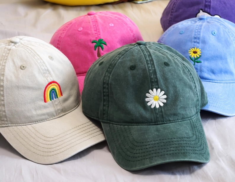 Puede incluir: Varias gorras de b&eacute;isbol en varios colores, incluyendo gris claro, rosa, azul y verde. Cada gorra presenta un dise&ntilde;o bordado, como un arco&iacute;ris, una palmera, una margarita y un girasol. Las gorras est&aacute;n hechas de un tejido suave y lavado.