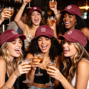 Puede incluir: Un grupo de personas con gorras de b&eacute;isbol granates con el texto "Cool Barre Club" en blanco, sosteniendo copas de bebidas y riendo. La escena tiene lugar en un bar, con un fondo oscuro.