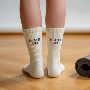 May include: A pair of off-white ribbed crew socks featuring the embroidered text "PILATES LIFE" in multicolored letters on the back of each sock. The socks are mid-calf length and have a vertical ribbed texture throughout the fabric.