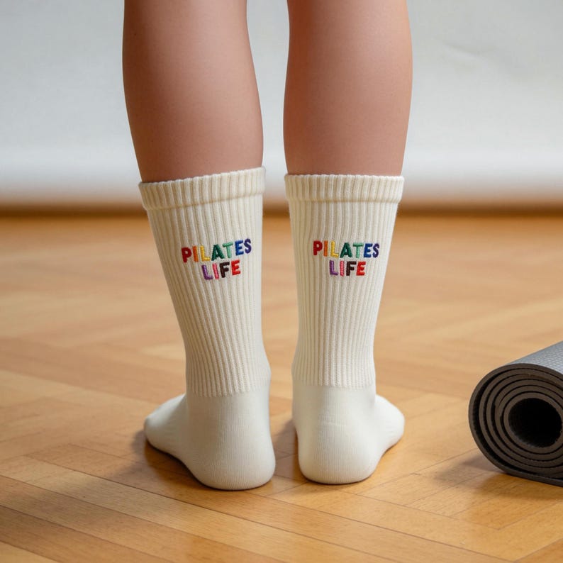 May include: A pair of off-white ribbed crew socks featuring the embroidered text "PILATES LIFE" in multicolored letters on the back of each sock. The socks are mid-calf length and have a vertical ribbed texture throughout the fabric.