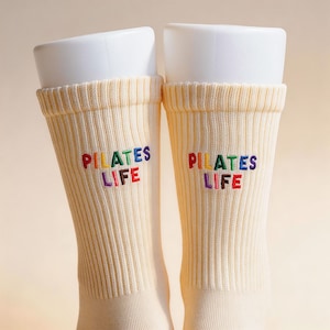 May include: A pair of cream-colored ribbed crew socks featuring the embroidered text "PILATES LIFE" in multi-colored letters. The letters are stitched in red, yellow, green, blue, and purple, creating a vibrant contrast against the light fabric.