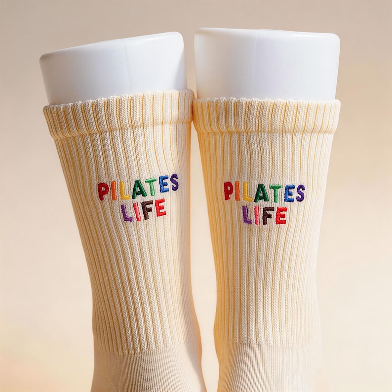 May include: A pair of cream-colored ribbed crew socks featuring the embroidered text "PILATES LIFE" in multi-colored letters. The letters are stitched in red, yellow, green, blue, and purple, creating a vibrant contrast against the light fabric.