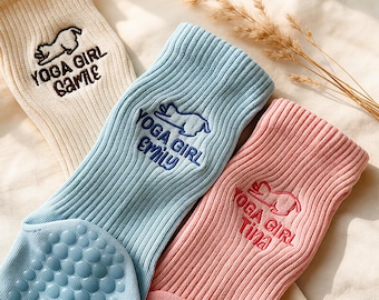 May include: Three ribbed crew socks in cream, light blue, and pink. Each sock features an embroidered cat silhouette and the text YOGA GIRL followed by a name: Camille, Emily, or Tina. The blue and pink socks have blue and pink grip dots.