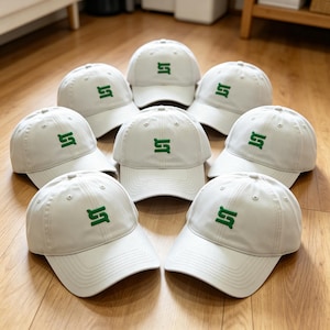 Puede incluir: Ocho gorras de b&eacute;isbol gris claro con un logotipo "S" verde bordado. Las gorras est&aacute;n dispuestas sobre una superficie de madera, mostrando su dise&ntilde;o y color.