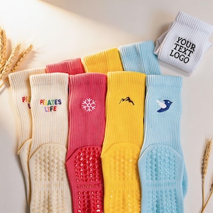 May include: A collection of ribbed crew socks in cream, pink, yellow, and light blue, featuring embroidered designs including a snowflake, mountain, bird, and the text "PILATES LIFE" and "YOUR TEXT LOGO". Each sock has a textured grip sole.