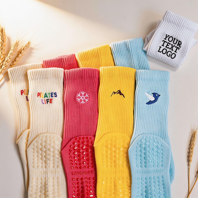 May include: A collection of ribbed crew socks in cream, pink, yellow, and light blue, featuring embroidered designs including a snowflake, mountain, bird, and the text "PILATES LIFE" and "YOUR TEXT LOGO". Each sock has a textured grip sole.