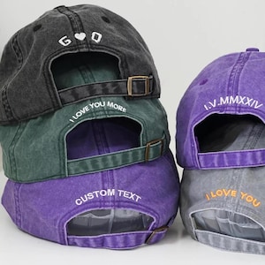 Puede incluir: Una pila de gorras de b&eacute;isbol en varios colores: carb&oacute;n, verde, morado y gris. Cada gorra tiene una visera curva y una correa ajustable. Las gorras presentan texto bordado, incluyendo "I LOVE YOU MORE" y "CUSTOM TEXT".