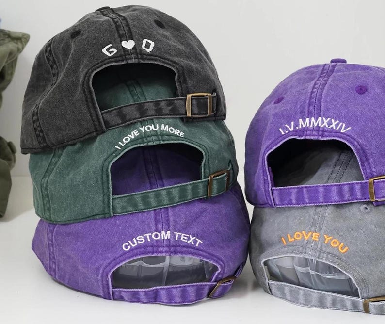 Puede incluir: Una pila de gorras de b&eacute;isbol en varios colores: carb&oacute;n, verde, morado y gris. Cada gorra tiene una visera curva y una correa ajustable. Las gorras presentan texto bordado, incluyendo "I LOVE YOU MORE" y "CUSTOM TEXT".