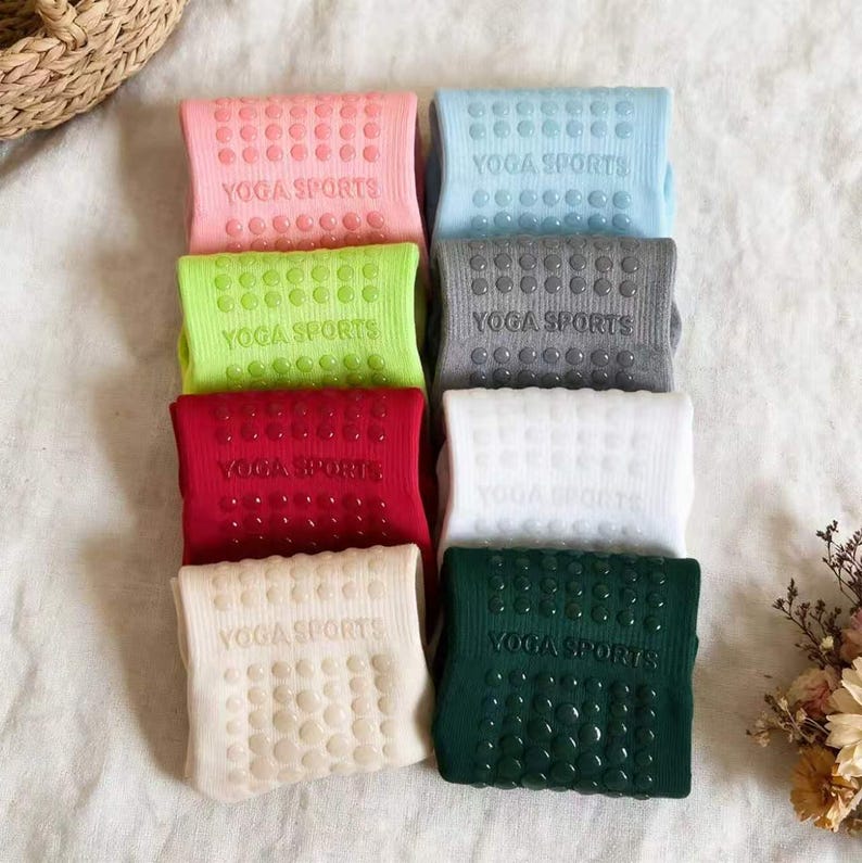 May include: A collection of eight folded socks in various colors including pink, light blue, lime green, gray, red, white, beige, and dark green. Each sock features a ribbed texture, the text YOGA SPORTS, and raised silicone grip dots.