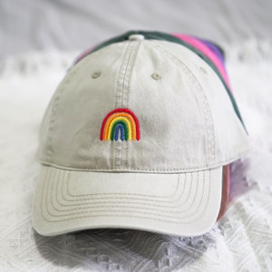 Può includere: Un berretto da baseball beige con un arcobaleno ricamato sul davanti.