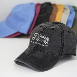 Puede incluir: Una gorra de b&eacute;isbol negra, desgastada, con la palabra "JESUS" bordada en blanco en la parte delantera. El texto debajo dice "YO SOY EL CAMINO, LA VERDAD, LA VIDA".