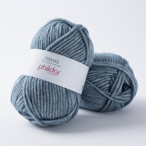 May include: Two balls of blue-grey yarn. The yarn is thick and appears to be made of a soft material. A white label wrapped around one ball reads "FRIMAS 100% mati&egrave;res naturelles phildar www.phildar.fr". The yarn is suitable for knitting or crochet projects.