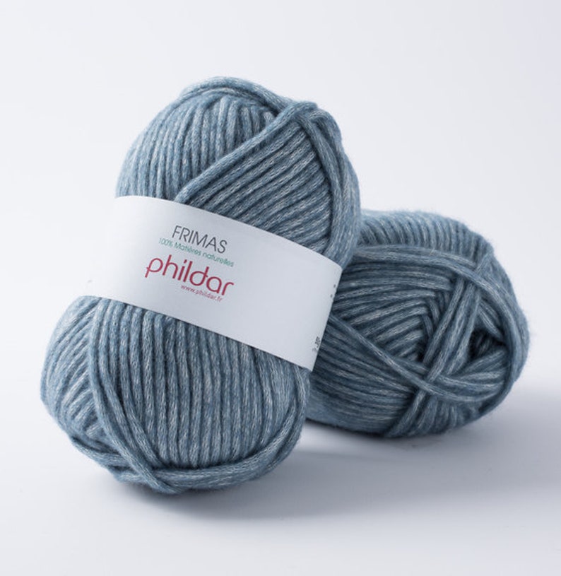 May include: Two balls of blue-grey yarn. The yarn is thick and appears to be made of a soft material. A white label wrapped around one ball reads "FRIMAS 100% mati&egrave;res naturelles phildar www.phildar.fr". The yarn is suitable for knitting or crochet projects.