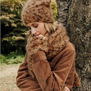 May include: A fuzzy, brown, leopard-print knit beanie and a matching scarf. The beanie has a soft, textured surface with a dark brown spotted pattern. The scarf is wrapped around the neck, featuring the same brown and black animal print.