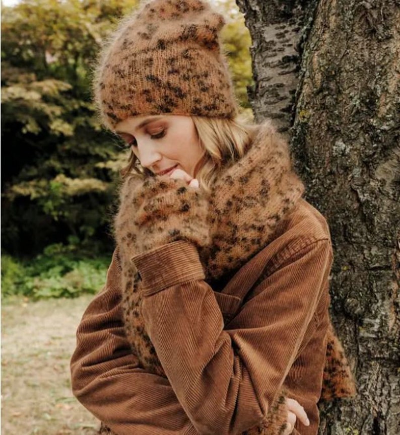 May include: A fuzzy, brown, leopard-print knit beanie and a matching scarf. The beanie has a soft, textured surface with a dark brown spotted pattern. The scarf is wrapped around the neck, featuring the same brown and black animal print.