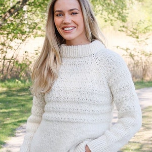 May include: A cream-colored long-sleeve sweater featuring a chunky knit texture, a high mock neck collar, and ribbed cuffs. The garment is crafted with a combination of smooth stockinette stitch sections and horizontal bands of textured knit patterns.