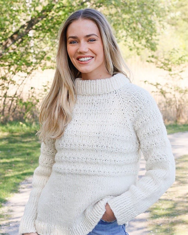 May include: A cream-colored long-sleeve sweater featuring a chunky knit texture, a high mock neck collar, and ribbed cuffs. The garment is crafted with a combination of smooth stockinette stitch sections and horizontal bands of textured knit patterns.