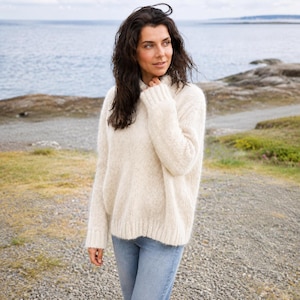 May include: A cream-colored, long-sleeved, fuzzy knit sweater with a high neckline and ribbed cuffs, paired with light blue denim jeans. The sweater features a soft, textured appearance with a loose fit and a straight hemline at the waist.
