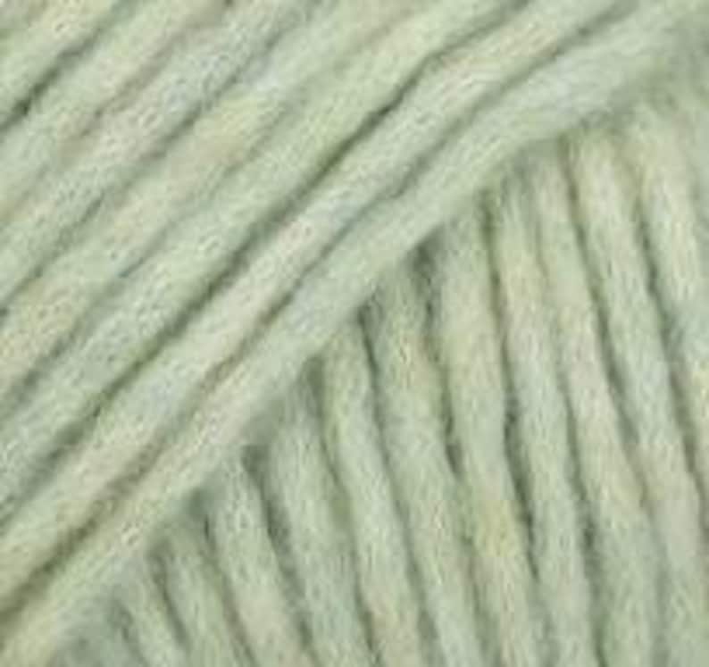 May include: A close-up view of light sage green yarn, showcasing the twisted fiber construction and soft, fuzzy texture of the strands. The yarn is arranged in parallel diagonal lines, highlighting the consistent ply and matte finish of the material.