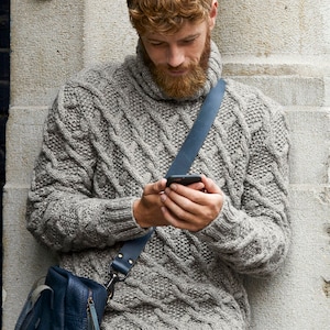May include: A gray cable-knit sweater with a turtleneck collar. The sweater has a textured pattern and is worn with a blue leather shoulder bag. The person is holding a smartphone.