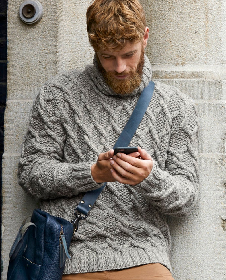 May include: A gray cable-knit sweater with a turtleneck collar. The sweater has a textured pattern and is worn with a blue leather shoulder bag. The person is holding a smartphone.