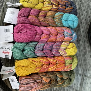Pode incluir: Uma coleção de oito novelos de fio em várias cores, incluindo amarelo, laranja, rosa, verde, azul e marrom. O fio está disposto em um padrão trançado e etiquetado como "Katia Edition Palette Hand Painted Yarn".