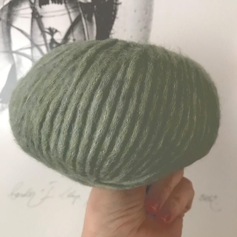 May include: A round, sage green ball of yarn featuring a distinct ribbed texture. The yarn has a soft, fuzzy appearance with visible fibers throughout the surface, creating a dense and uniform look across the entire spherical shape of the object.