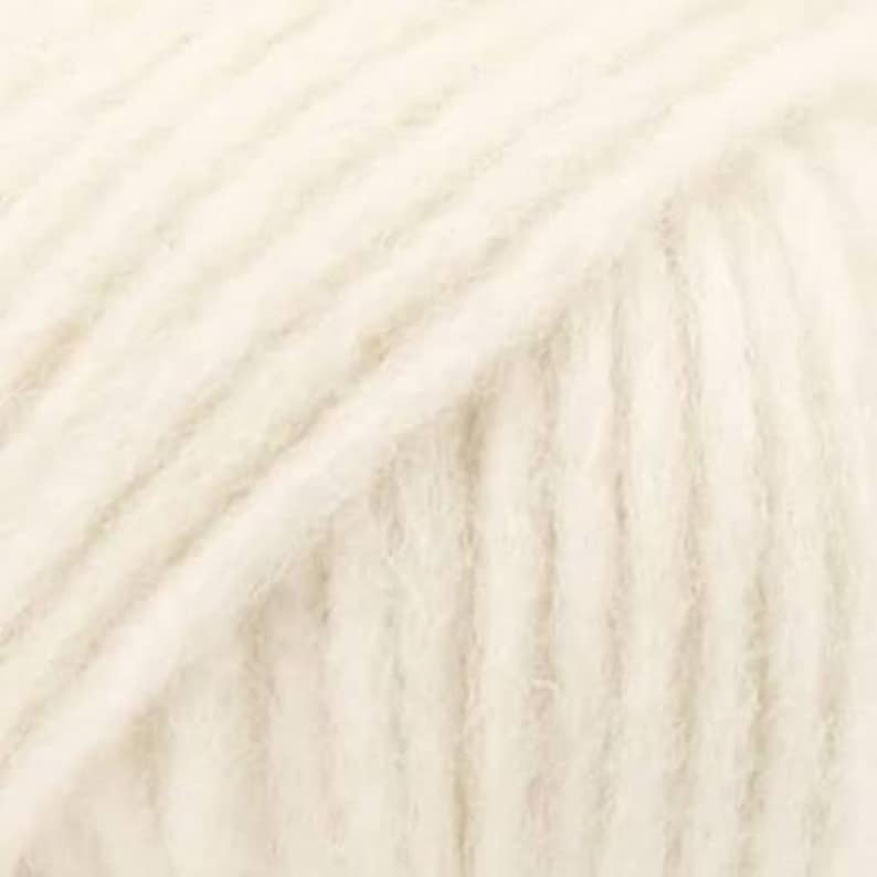 May include: A skein of off-white yarn featuring a soft, fuzzy texture with visible twisted plies. The yarn is wound in a dense, uniform pattern, showcasing a light cream color throughout the entire surface of the fiber bundle.