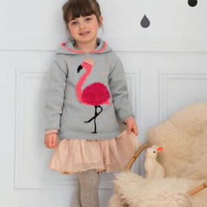 Peut inclure: Une jeune fille portant un sweat &agrave; capuche gris avec un motif de flamant rose. Le sweat-shirt a une bordure rose autour de la capuche. La fille porte une jupe en tulle rose clair et des collants gris.