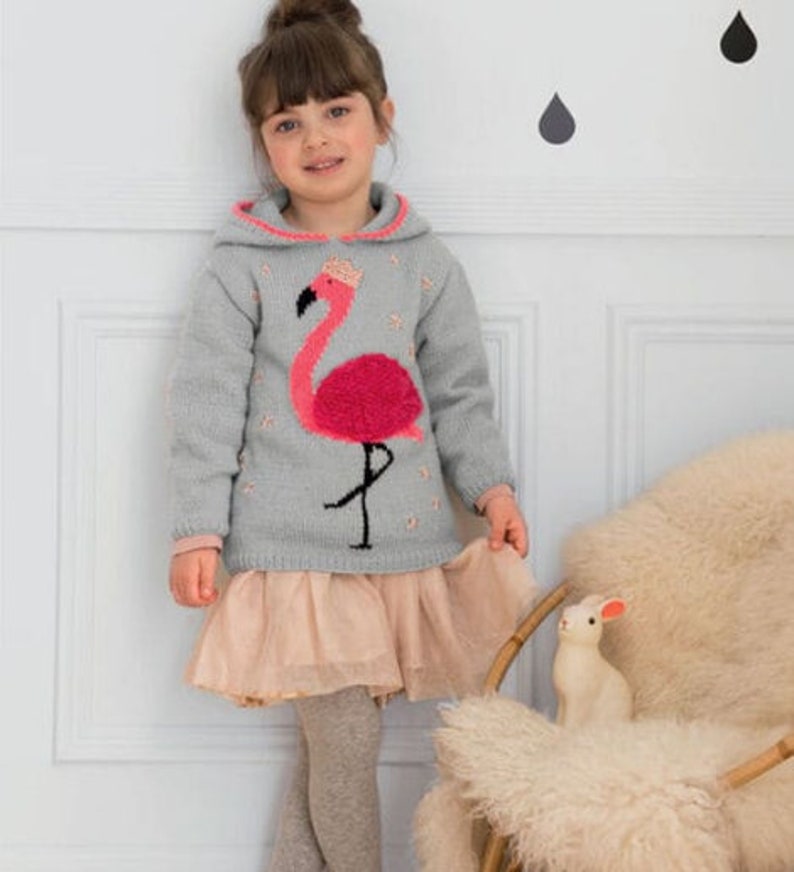 Peut inclure: Une jeune fille portant un sweat &agrave; capuche gris avec un motif de flamant rose. Le sweat-shirt a une bordure rose autour de la capuche. La fille porte une jupe en tulle rose clair et des collants gris.