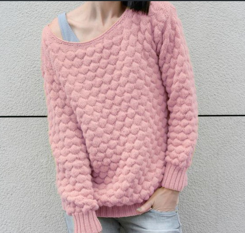 May include: A pink knitted sweater with a textured, heart-shaped pattern. The sweater has a wide, off-the-shoulder neckline and long sleeves.