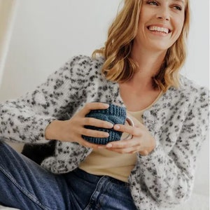 May include: A fuzzy white cardigan featuring a black leopard print pattern, worn over a plain beige tank top and blue denim jeans. The cardigan has long sleeves and a button-down front, paired with a dark blue knitted mug cozy held in the hands.