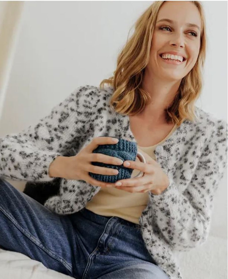 May include: A fuzzy white cardigan featuring a black leopard print pattern, worn over a plain beige tank top and blue denim jeans. The cardigan has long sleeves and a button-down front, paired with a dark blue knitted mug cozy held in the hands.