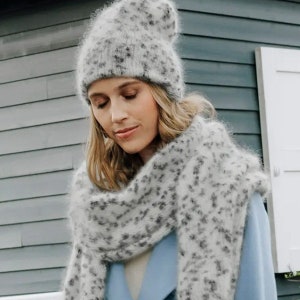 May include: A fuzzy, light gray beanie hat and a matching long scarf, both featuring a speckled pattern of small dark gray spots. The knit accessories have a soft, textured appearance and are worn over a solid light blue coat with a lapel collar.