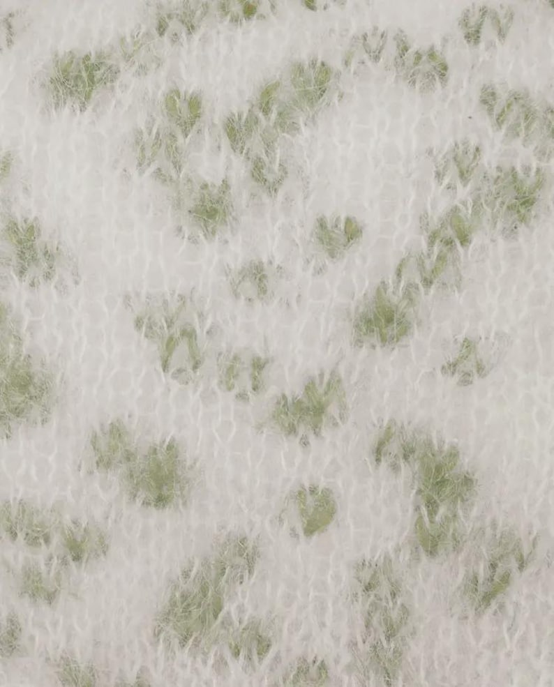 May include: A close-up view of a soft, fuzzy knit fabric featuring a white base color with an irregular, scattered pattern of light sage green spots. The material has a distinct, slightly brushed, and textured appearance with visible yarn loops.