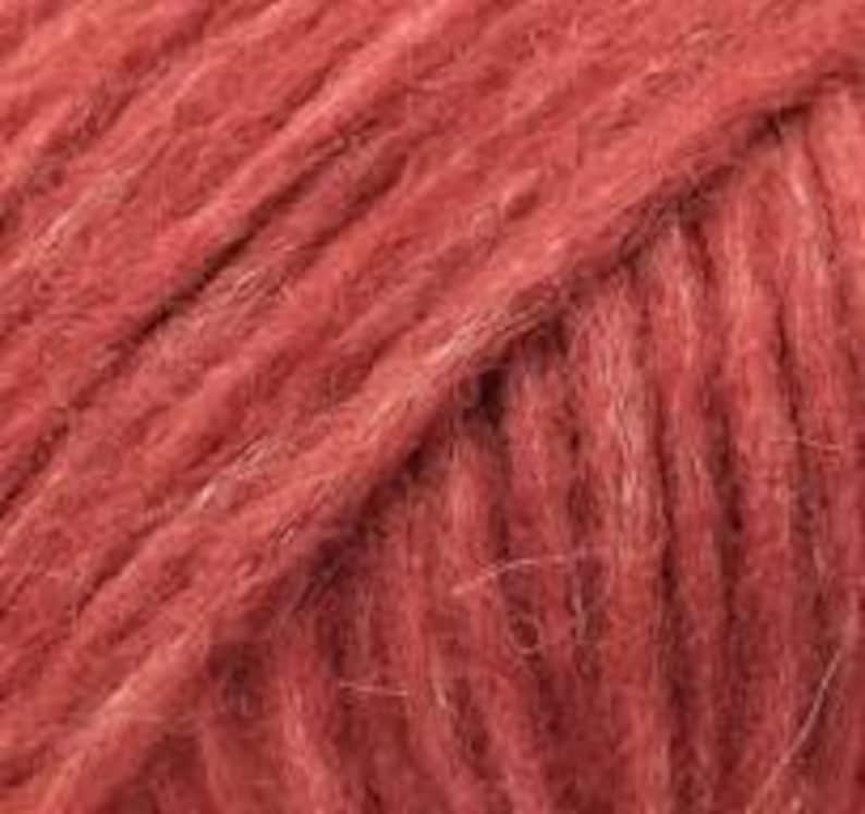 May include: A close-up view of a skein of yarn in a deep, muted terracotta red color. The texture is fuzzy and fibrous, showing the twisted strands of the yarn running diagonally across the frame, highlighting the soft, matte finish of the material.