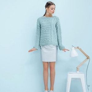 May include: A light blue cable-knit sweater with a crew neck. The sweater has a textured pattern and long sleeves. The model is wearing a light gray skirt. A white lamp with a wooden frame is on a white stool.