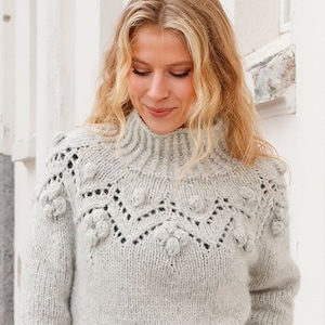 May include: A light gray long-sleeve sweater featuring a high ribbed collar and a textured yoke with intricate knit patterns, including small raised bobble details and openwork stitches across the chest and shoulders in a soft wool blend.