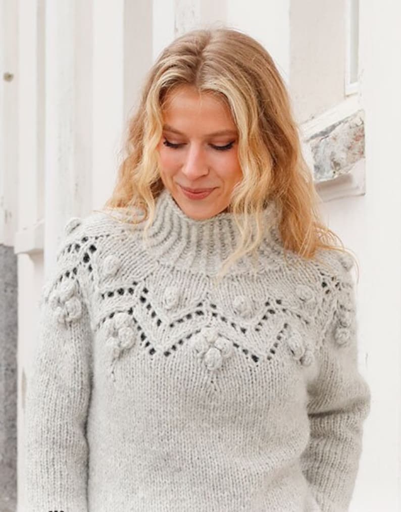 May include: A light gray long-sleeve sweater featuring a high ribbed collar and a textured yoke with intricate knit patterns, including small raised bobble details and openwork stitches across the chest and shoulders in a soft wool blend.
