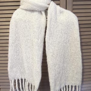 Puede incluir: Bufanda de punto blanco roto con borde de flecos. La bufanda está hecha de un hilo grueso y texturizado. La bufanda está sobre una superficie de madera. Un accesorio de invierno.
