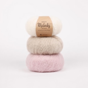 May include: A stack of three balls of fuzzy yarn in white, beige, and light pink. The top ball features a tan paper label with the text DROPS Melody, Uni Colour, and A luxurious mix of wool and alpaca printed in dark brown ink on the front.
