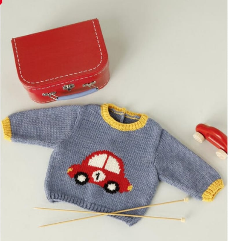 Peut inclure: Un pull en tricot bleu avec une bordure jaune et un motif de voiture rouge sur le devant. Le pull est pos&eacute; sur une surface blanche avec deux aiguilles &agrave; tricoter en bois.