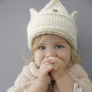May include: A young child wearing a white knitted crown hat with a wooden button on top. The child is smiling and has their hands clasped in front of their face.