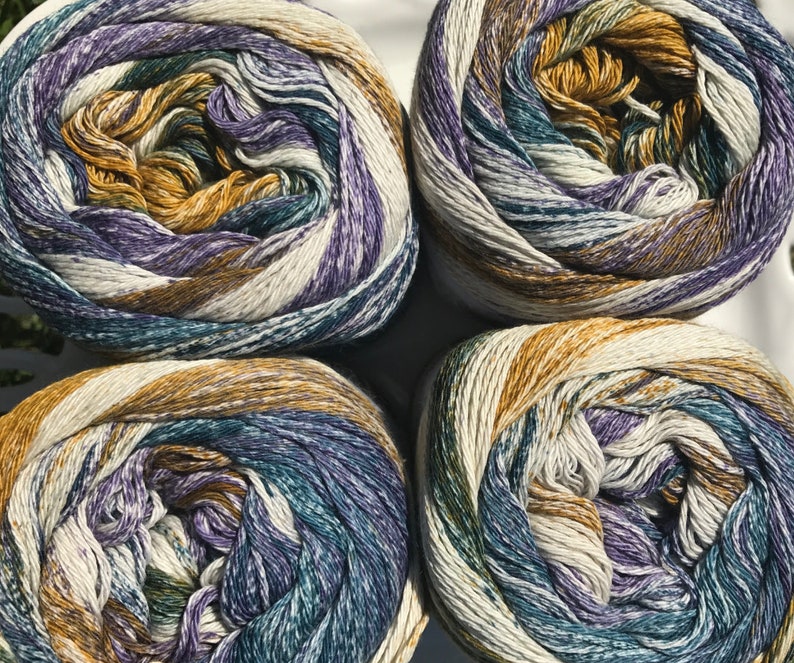 May include: Four balls of yarn in a white, purple, teal, and brown color scheme. The yarn is arranged in a circle on a white surface.