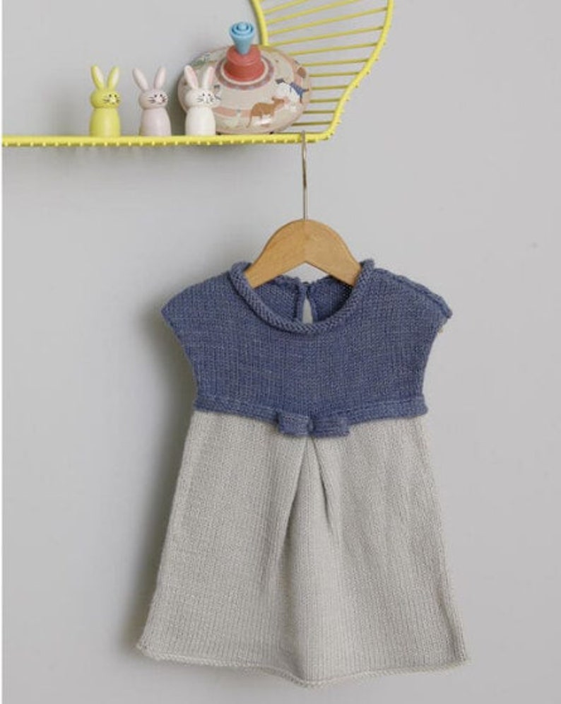 Peut inclure: Une robe en tricot bleu et gris pour b&eacute;b&eacute; avec un n&oelig;ud &agrave; la taille. La robe est suspendue &agrave; un cintre en bois.