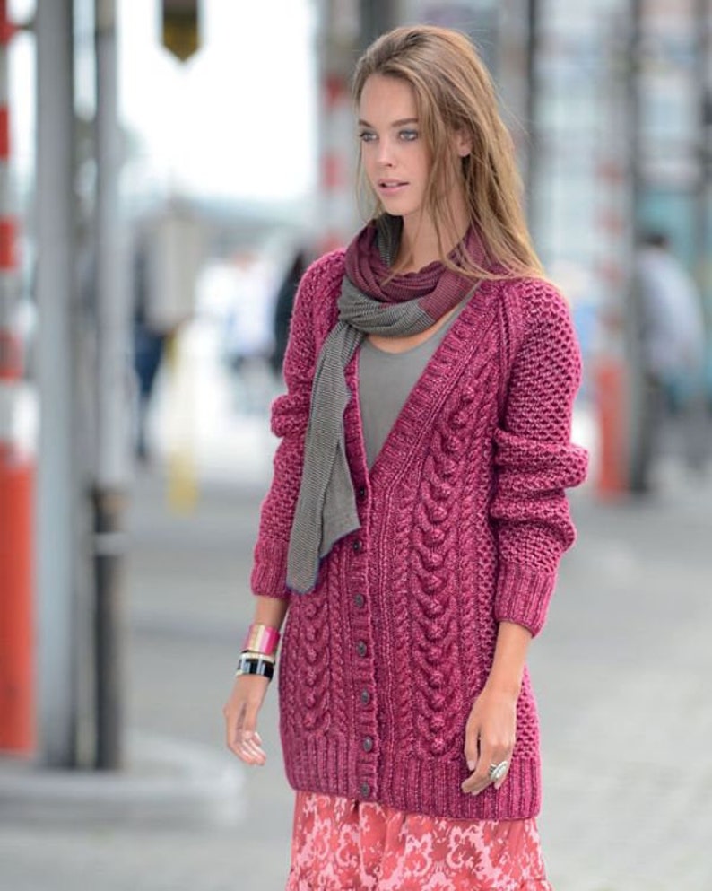 May include: A woman wearing a long, button-down, magenta cable-knit cardigan with a gray scarf. The cardigan has a textured pattern and long sleeves. She is also wearing a pink and red patterned skirt and a bracelet.