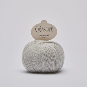 May include: A round ball of light gray yarn with a textured, soft appearance. A beige paper tag is attached to the top, featuring the text Cardiff Cashmere and a small goat icon, with the word small printed below the brand name.