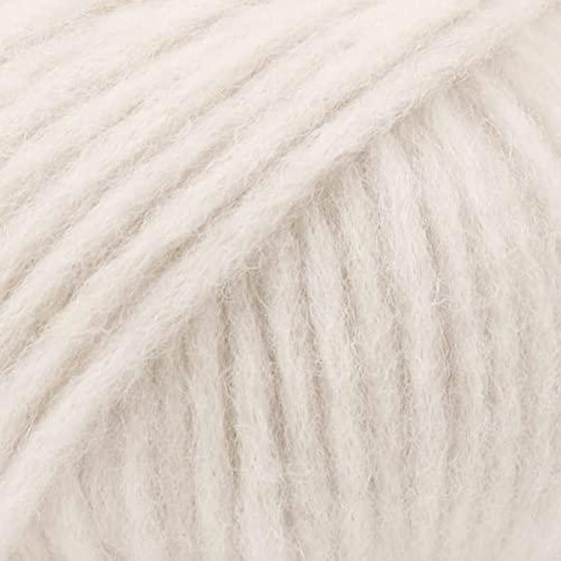 May include: A skein of off-white yarn featuring a soft, fuzzy texture with visible individual strands twisted together. The fibers have a light, airy appearance with a consistent pale cream color throughout the entire surface of the yarn ball.