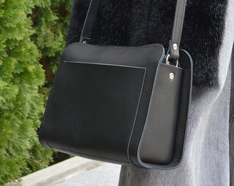 Può includere: Una borsa a tracolla nera strutturata con finitura opaca liscia, caratterizzata da una silhouette rettangolare, una sottile tracolla regolabile con fibbia in metallo argentato e soffietti laterali visibili fissati con piccoli rivetti in metallo argentato.